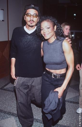 Janet Jackson smiling while holding a gray jacket and René Elizondo Jr. is looking seriously with a mustache and beard. Janet with black curly hair is wearing a gray sleeveless blouse and black pants with a black belt. René is wearing a sunglasses, a black cap, gray pants, and a white shirt under a black long sleeve shirt