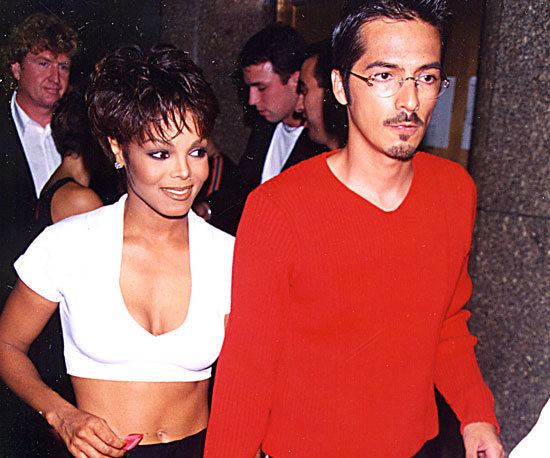 Janet Jackson smiling and René Elizondo Jr. is looking seriously with a mustache and beard, and there are some people behind them. Janet is wearing a white crop top with a low-cut neckline that exposes her cleavage while René is wearing eyeglasses and red long sleeve shirt