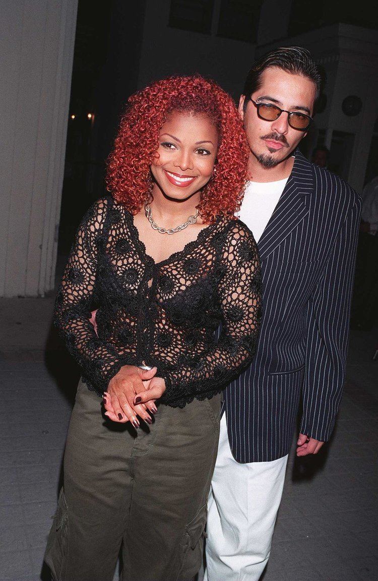 Janet Jackson smiling and René Elizondo Jr. is looking seriously with a mustache and beard. Janet with red curly hair is wearing moss green pants, a necklace, earrings, and a black brassiere under a black long sleeve crochet blouse. René is wearing sunglasses, white pants, and a white shirt under a blue striped coat