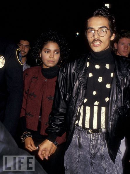 Janet Jackson and René Elizondo Jr. attend the Eddie Murphy's Comedy Tour at The Forum in Inglewood, California. Janet with a serious face and black curly hair is wearing hoop earrings, a bracelet on her right wrist and a watch on her left wrist, and a black long sleeve blouse under a maroon cardigan. René with a serious face, mustache, and mouth is open while wearing denim pants and a black and white turtleneck shirt under a black leather jacket
