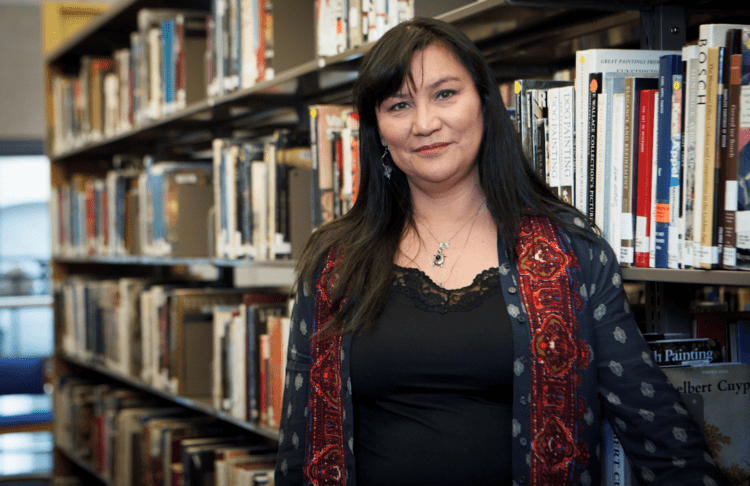 Renae Morriseau smiling and standing beside the bookshelves and wearing a black shirt under a blue and red cardigan