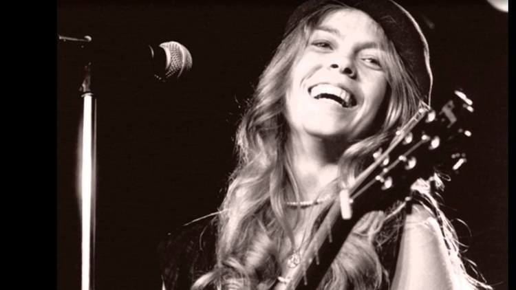 Renée Fladen Kamm happily singing and playing a guitar with her wavy hair and wearing a hat.