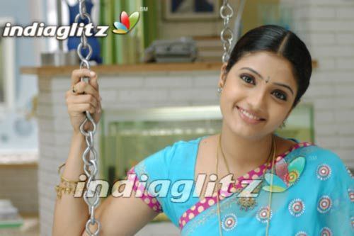 Renuka Menon smiling while wearing a sky-blue and pink blouse, earrings, necklace, and bracelet