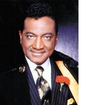Reverend Ike smiling and wearing a white sleeve, navy blue suit, and a black coat with an orange boutonniere