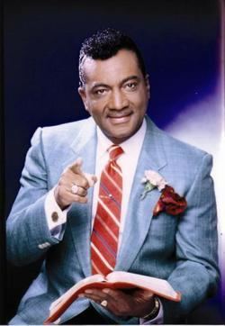 Reverend Ike smiling, holding a book and pointing his finger while wearing white long sleeves, gray coat, and a red-striped necktie