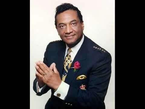 Reverend Ike smiling and pressing his fingers together while wearing a  white suit and navy blue coat