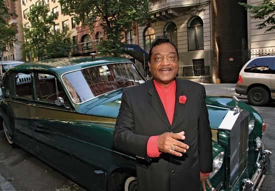 Reverend Ike smiling while standing at the side of a green car with a curly black hair and wearing pink sleeves and black tuxedo