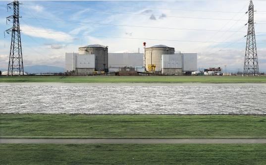 The Rhine river flows horizontally across the field between flat green fields under an overcast sky in front of a factory building and transmission towers.