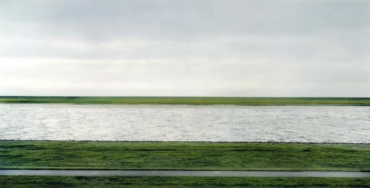 A river flows horizontally across the field between flat green fields under an overcast sky.