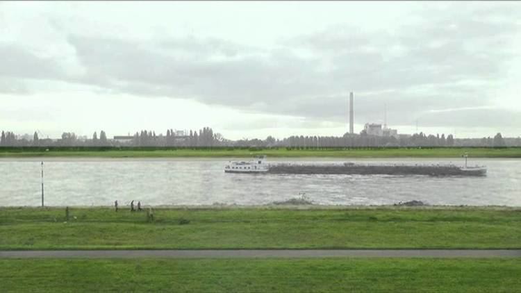 The Rhine river with boats flows horizontally across the field between flat green fields with people walking their dogs under an overcast sky.
