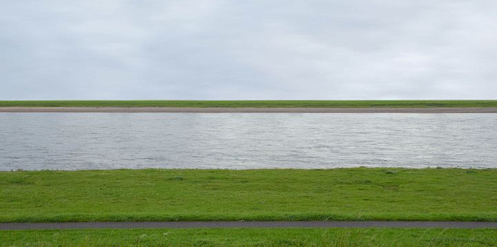 The Rhine river flows horizontally across the field between flat green fields under an overcast sky.