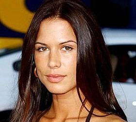 Rhona Mitra looking at something with a neutral face, brown eyes and long brown hair, gold loop earring, and a white car in the background, wearing a spaghetti-strapped top.