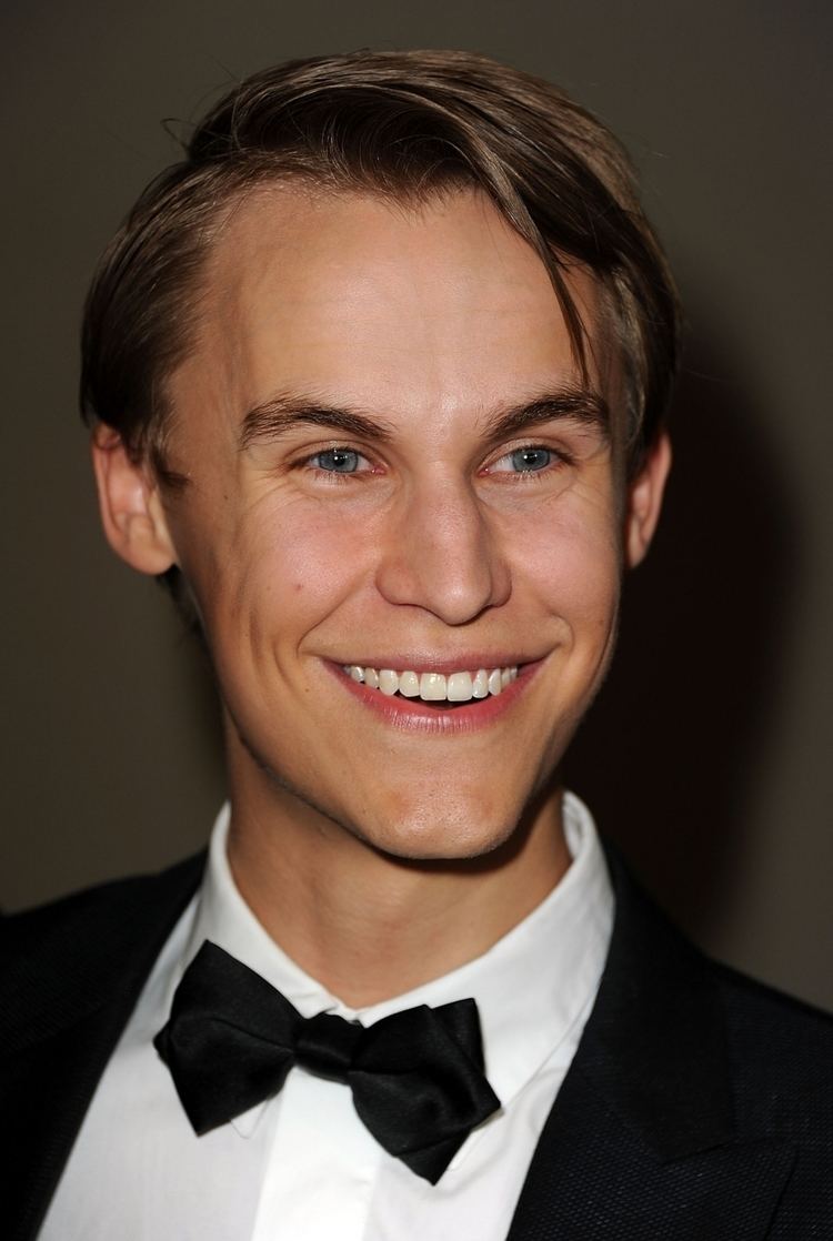 Rhys Wakefield smiling with his black hair wearing a white polo long sleeve with a black bowtie under a black coat