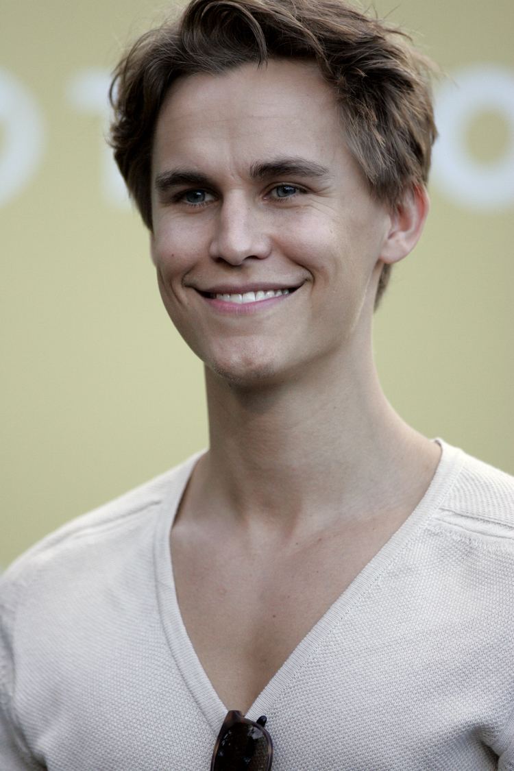 Rhys Wakefield smiling with his blonde hair wearing a V-neck white shirt with a black sunglasses on it