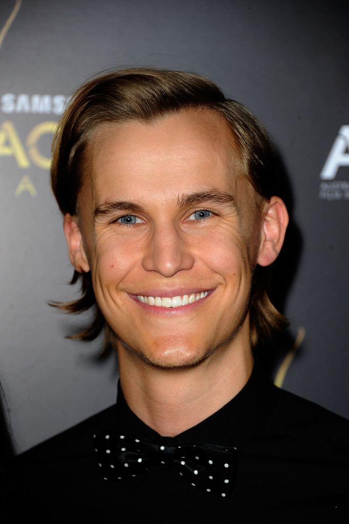 Rhys Wakefield smiling with his blonde hair wearing black long sleeves with a black and white polka dots bowtie