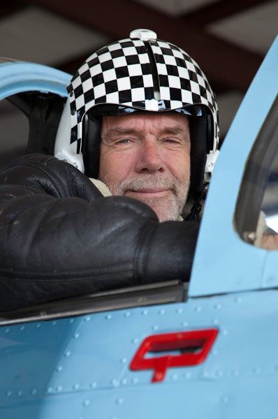 Richard Bach smiling, wearing a black jacket, a white and black helmet inside a car.