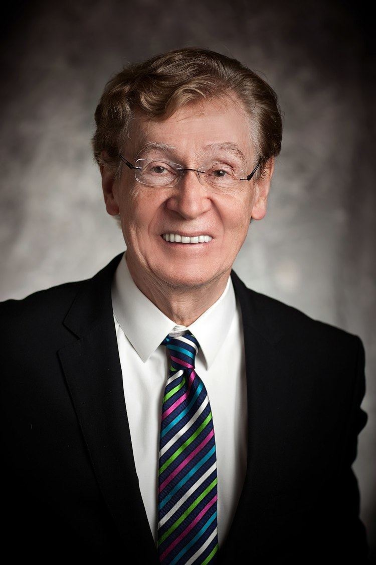 Richard Driehaus smiling, wearing eyeglasses, a black suit, white long sleeves, and a colorful tie.