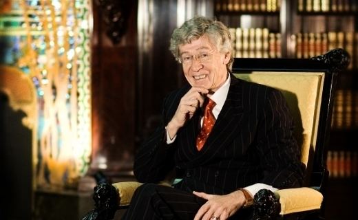 Richard Driehaus smiling while sitting on a chair and wearing a black suit and a red tie.