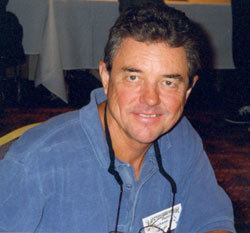 Richard Eyer smiling and sitting on a chair with a black strapped-eyeglasses hanging around his neck and a badge card pinned on his shirt at the Memphis Film Festival in 1998, with a white cloth in the background, wearing a blue collared-long sleeve.