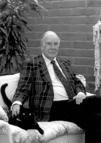 Richard Hamming smiling while sitting on the couch with his cat and wearing a checkered coat, white long sleeves, necktie, and pants