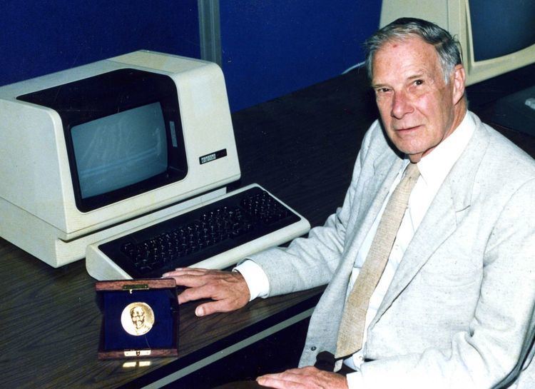 Richard Hamming smiling while a vintage computer at his side and he is wearing a gray coat, white long sleeves, and beige necktie