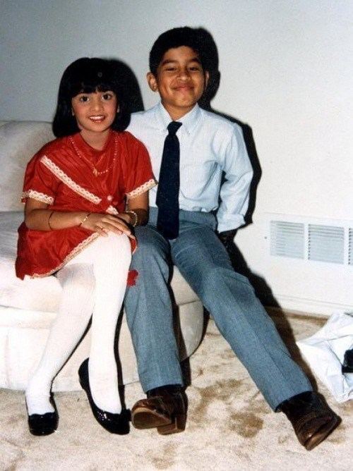 Young Richard Rishi smiling, wearing long sleeves and a black tie with a little girl wearing a red dress, long white socks, and black shoes.