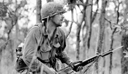 Retired Army Reserve Col. Rick Rescorla at the Ia Drang battle while holding a rifle, carrying a bag, and wearing a helmet and long sleeves