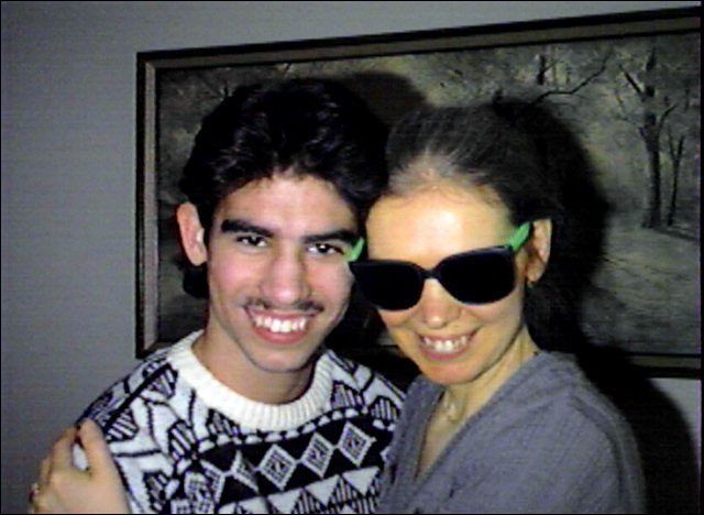 Ricky Rodriguez smiling and wearing a black and white sweatshirt while the woman beside him wearing a gray blouse and black shades