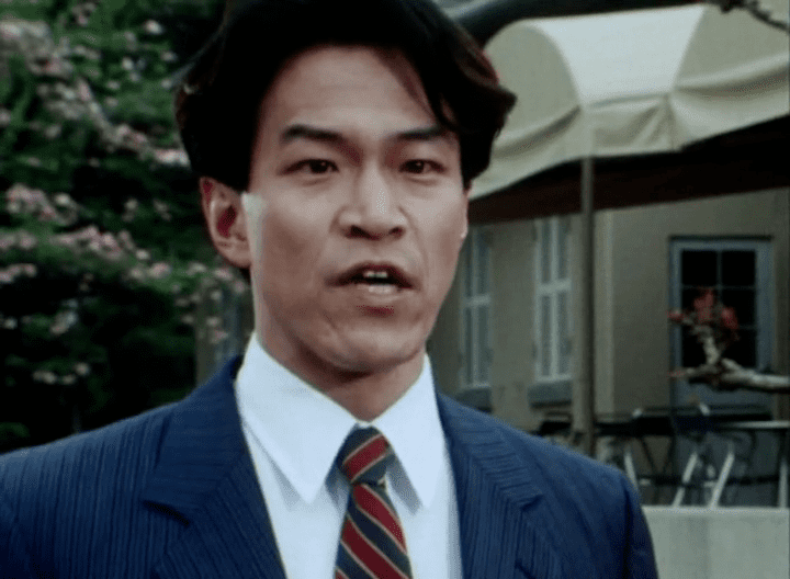 Rikiya Koyama talking to someone while wearing a blue coat, white long sleeves, and red and blue striped necktie