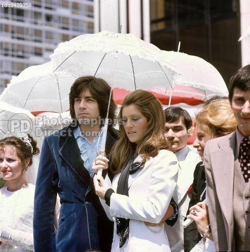 Ringo and Sheila with serious faces, while Sheila holding a white umbrella and surrounded by people. Ringo wearing a blue coat over a light blue shirt while Shiela wearing a white long sleeve dress with a black scarf.