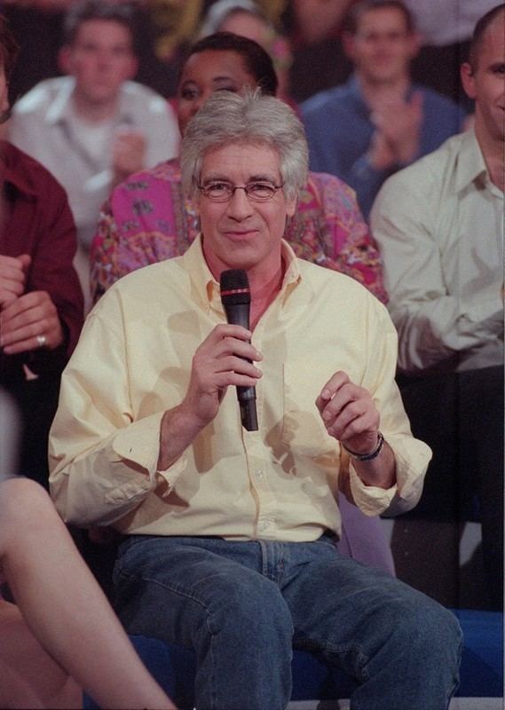 Ringo is talking and holding a microphone while sitting on a chair with an audience on his back, with white hair, wearing a yellow polo shirt and blue jeans.
