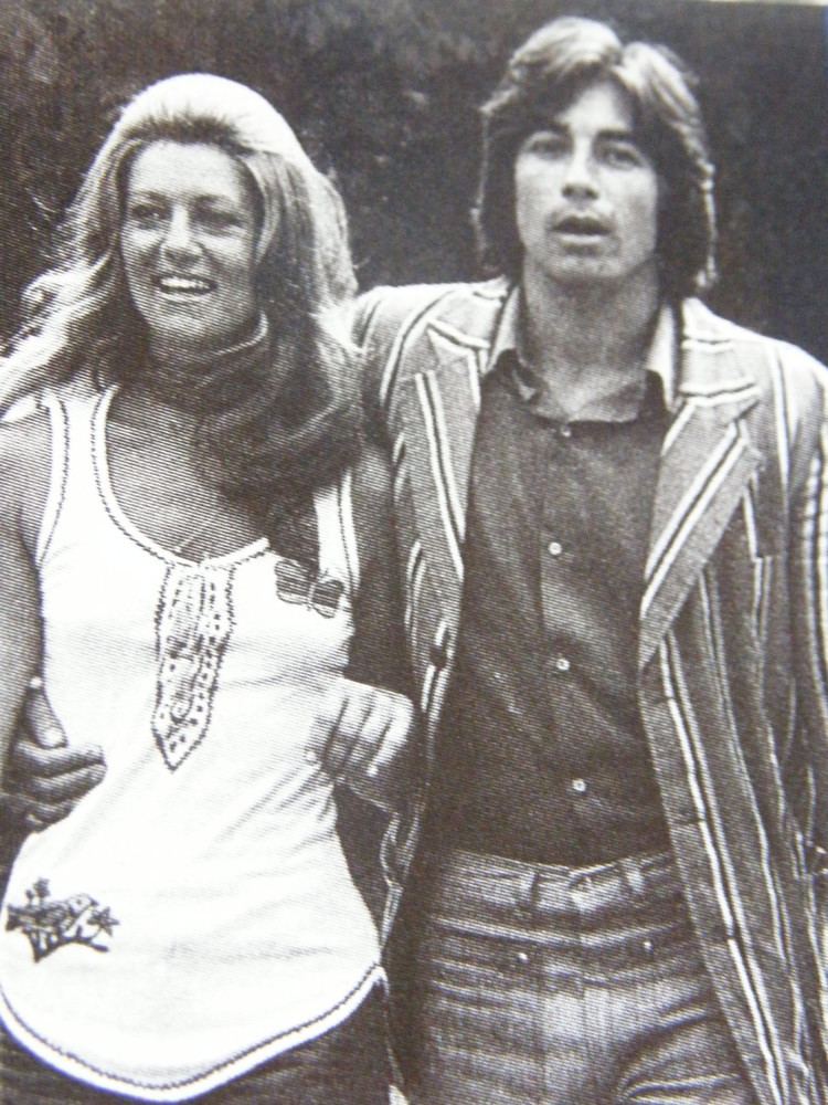 Shiela smiling while Ringo holding her waist. Shiela with wavy blonde hair and wearing a white sleeveless top while Ringo wearing a striped coat over a black polo shirt, and jeans.