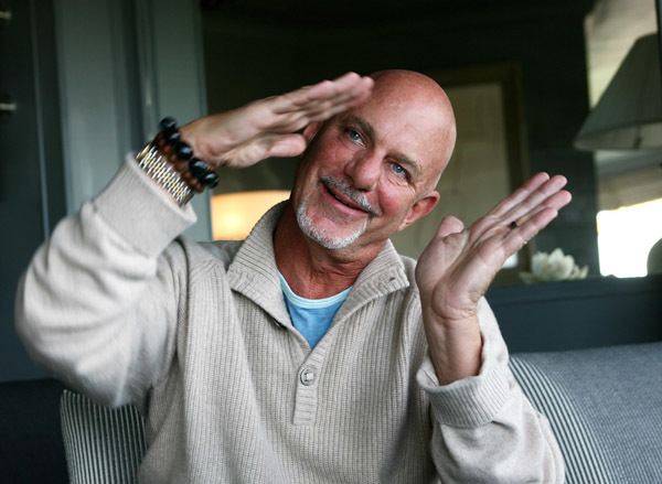 Rob Cohen smiling and tilting his head on the right side together with his hand while wearing a gray knitted sweater with collar and blue shirt underneath