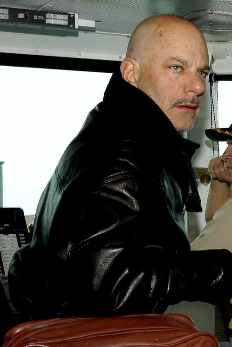 Rob Cohen looking afar while wearing a black leather jacket and beside him is a brown leather bag