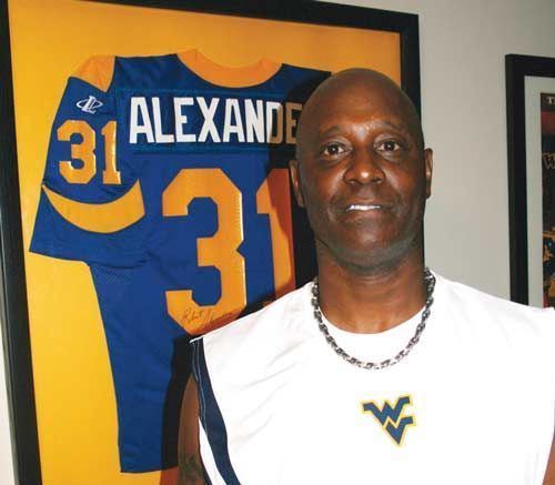 Robert Alexander is smiling, bald, a frame of his uniform with his name at the back, with a print number 31 at the center and on the right side, and his name “ALEXANDER”, he is wearing a black necklace and a white shirt with a print “WV”.