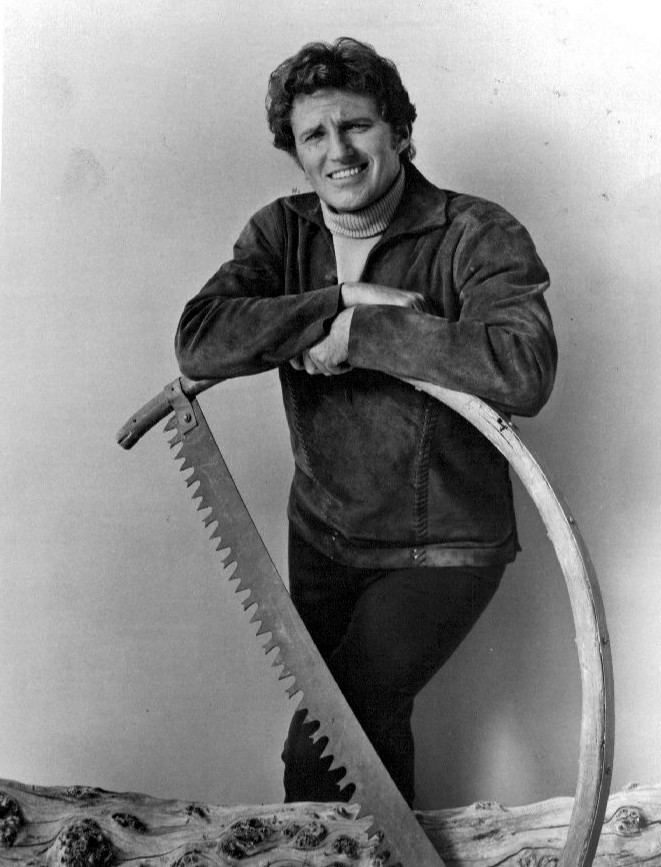 Robert Brown smiling and leaning on a big saw while wearing a jacket, turtle neck and pants