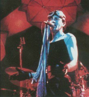 Robert Calvert is singing, standing in front of the microphone, arms bent palm up open, has black hair wearing motorcycle goggles, black gloves, black sleeveless top and black pants behind him is a drumset.