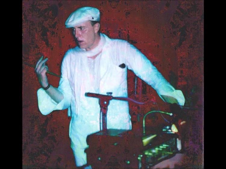 Robert Calvert, a musician, serious, standing, right hand holding a drumstick and left hand on a table with an amplifier set, has black hair wearing a white Newsboy hat, white scarf, black bracelet, white long sleeves, and white pants.