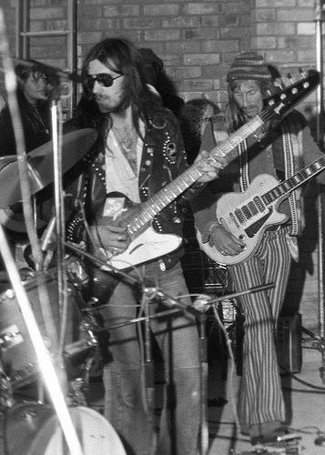 On the left is Robert Calvert, standing while playing with his guitar, looking at the drums, has long black hair and beard and a mustache, wearing a v cut white, denim pants and black printed jacket, with audience at the back, at the Right is Lemmy Kilmister, standing right foot forward, playing his white round guitar, has long black hair a mustache wearing a bonnet striped pants, and a long sleeve printed jacket.