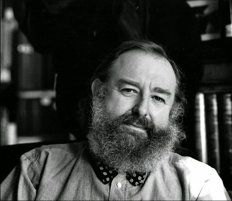 Michael Moorcock, a musician, is serious, sitting, has a long mustache and faded gray beard, black hair, wearing a polo and printed bowtie.