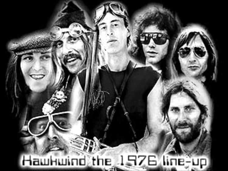 The Hawkwind 1976 lineup On the top left is Steve Peregrin, smiling, with long hair, wearing a newsboy cap, 2nd from left is Dave Brock, smiling, with a mustache, long black hair, wearing a pointy hat with goggles, 3rd from left, Robert Calvert, serious, standing, holding his guitar, wearing a scarf over his head with motorcycle goggles, 4th from left, a man is serious, with curly long hair, wearing black shades. On the left bottom is Arthur Brown, smiling, with long hair, wearing shades, at the bottom right, Jim Capaldi is smiling, has black wavy hair, mustache, and beard, wearing a polo,