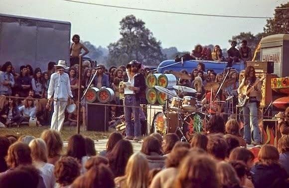 At the Windsor Free Festival in 1973, in the middle is a band with four people, and around the stage was the audience. From left, a man standing with a white tamborine, wearing a white hat, a white long sleeve and white pants, 2nd from left, a man standing, has a beard and mustache, wearing a black hat, white shirt, black jacket and denim pants, 3rd from left, a man is sitting playing drums, wearing a red shirt, at the right is a man standing, playing with his guitar, has long curly hair, wearing a yellow brown long sleeve and denim pants with a black belt.