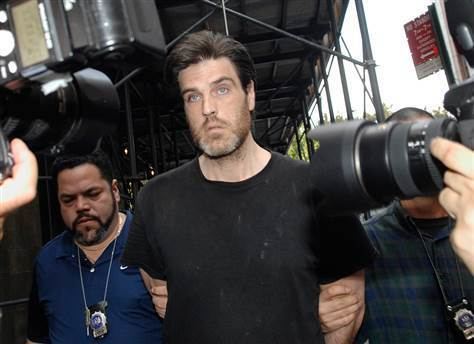 Robert Chambers and the two men beside him are looking at something while people are taking pictures of them, with serious faces, a mustache, and beards. Robert is wearing a black t-shirt, the man on his left is wearing a blue t-shirt and police id, and the man on his right is wearing a blue and green polo and police id