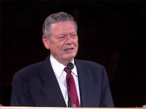 Robert E. Wells delivering a speech in front of a microphone having gray hair with a black background, wearing a white shirt under a dark blue coat and a red necktie