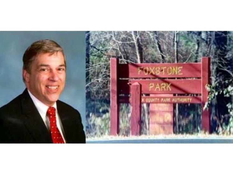 On the left, Robert Hanssen with a big smile and wearing a black coat, white long sleeve, and red necktie with cream and black pattern while on the right, is the Foxstone Park's signage