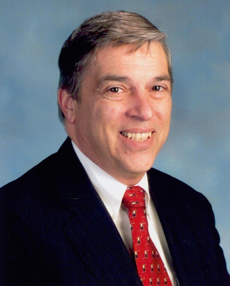 Robert Hanssen with a big smile while wearing a black coat, white long sleeve, and red necktie with cream and black pattern