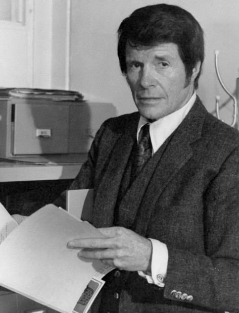 Robert Horton looking serious and holding a folder with a filing cabinet in the background, wearing a shirt under a vest, a coat and a necktie