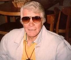 Robert Horton looking serious has white hair with a table and a basket and chairs in the background, he has a necklace and sunglasses-wearing a yellow shirt under a white coat