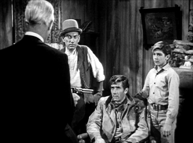 Young Robert L. Crawford Jr. standing beside Robert Fuller with a serious face while looking at a man holding a gun in a scene from Laramie, a 1559 American TV series.