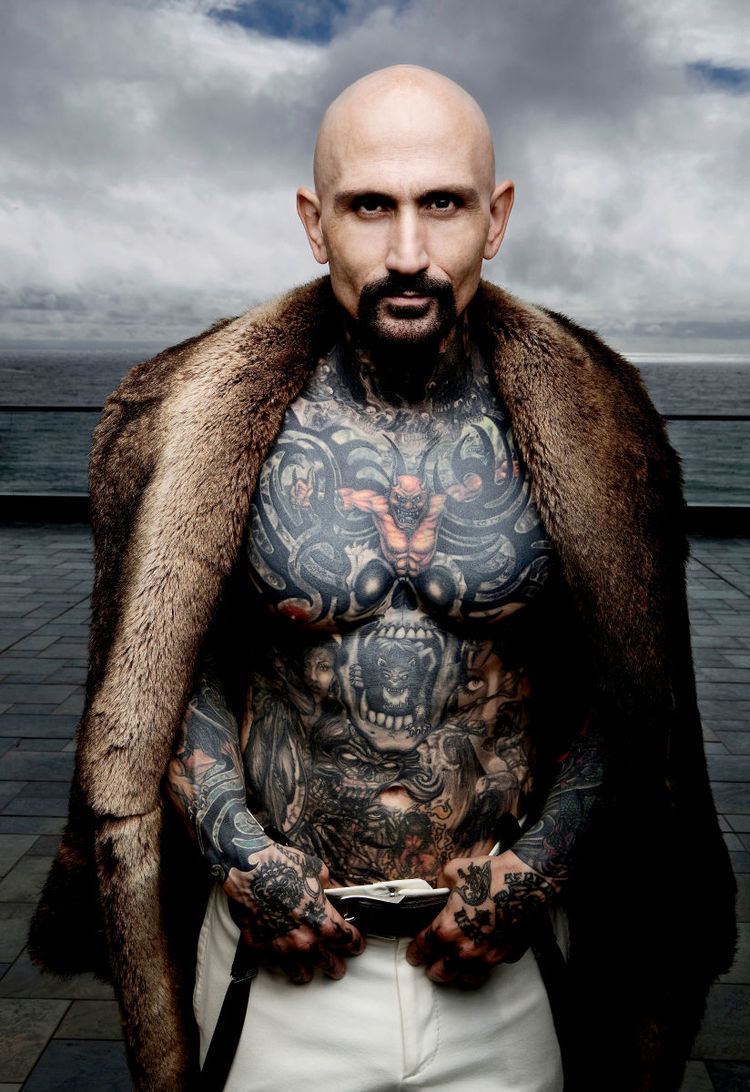 Robert LaSardo with a mustache, beard, and tattoos on his body while wearing a brown fur coat, black belt, and white pants